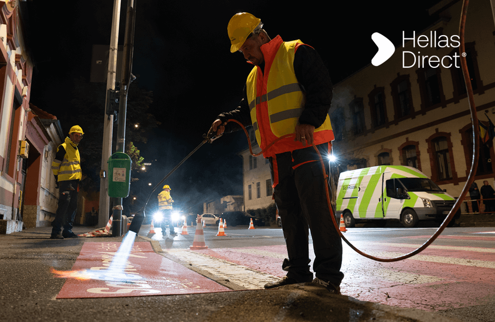 man in yellow vest paints crosswalk outside a school in the night. Written text: Hellas Direct